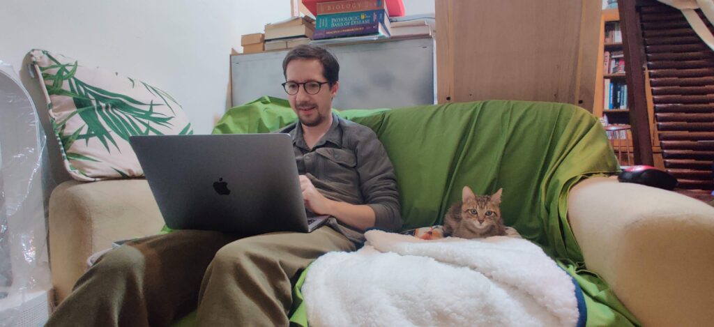 Me and our cat sitting on a sofa, the cat looking at the camera, and I'm working on my laptop.